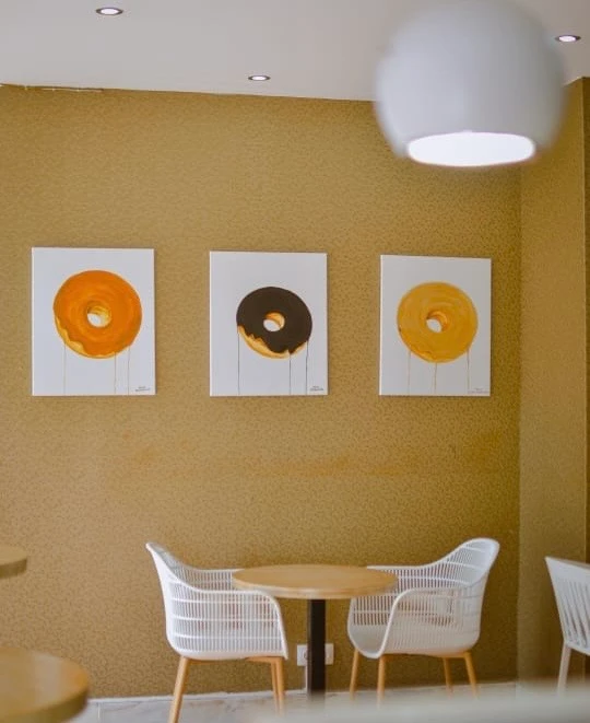 Interior da Kêro Donuts, com três quadros de donuts sobre uma mesa e cadeiras brancas.
